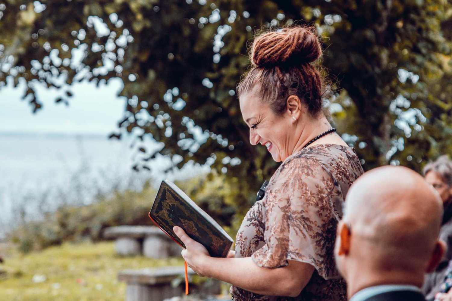 Priesterin erklärt, was ein Ritual ist, mit einem symbolgeschmückten Buch in der Hand unter freiem Himmel vor einem Baum