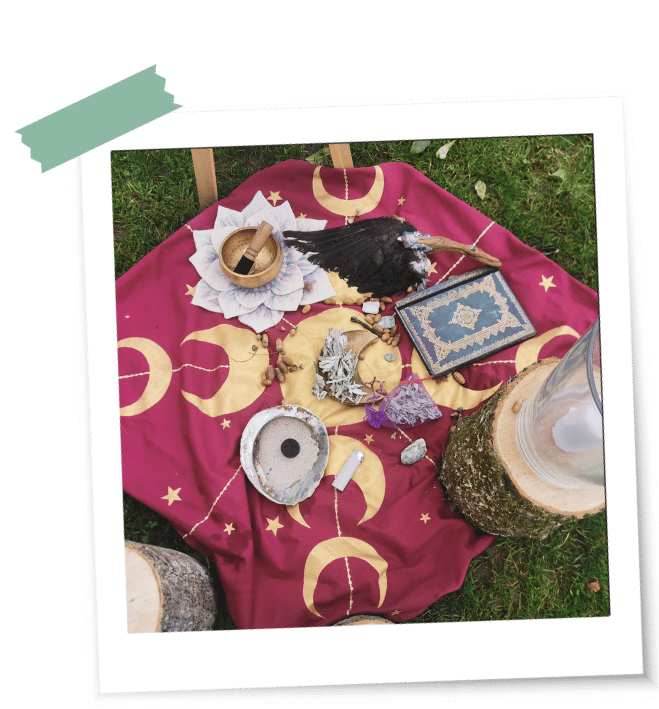 Was ist ein Ritual LaLuna-Ritualtuch Ein rotes LaLuna-Ritualtuch mit goldenen Mondphasen liegt auf einer Wiese mit Muscheln, einem Buch, Räucherfächer und Räucherwerk bei einem Ritual